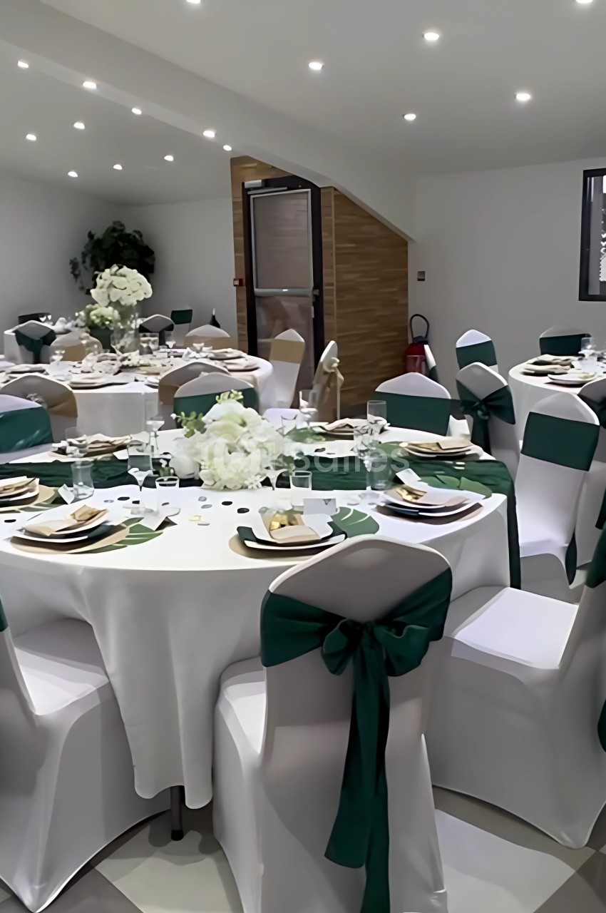 Salle décorée pour un événement, avec tables rondes dressées, nappes blanches et chaises ornées de nœuds verts.