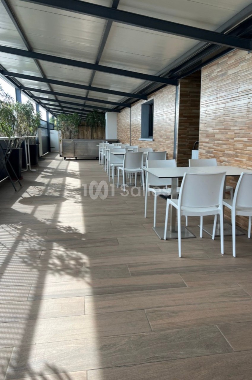 Terrasse couverte avec sol en bois, tables blanches alignées et murs en briques claires, éclairée par la lumière naturelle.