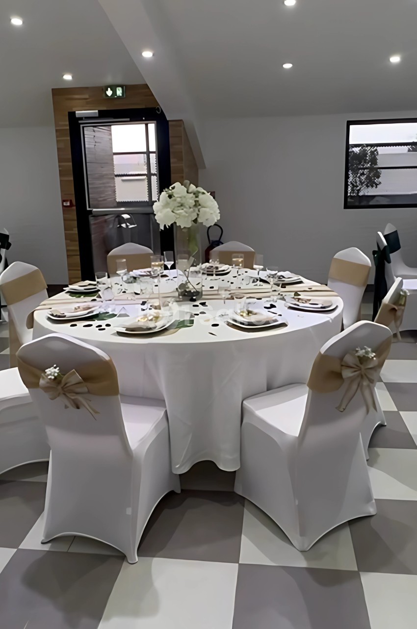 Table ronde décorée pour un événement, avec nappes blanches, chaises ornées de nœuds beige et centre de table floral.
