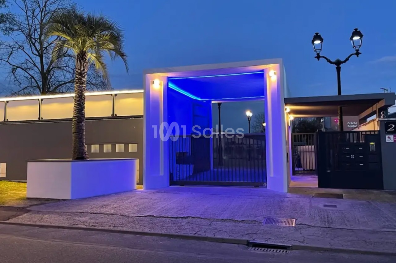 Entrée moderne illuminée en bleu avec une palmeraie et des lampadaires, devant un portail noir et un mur gris.
