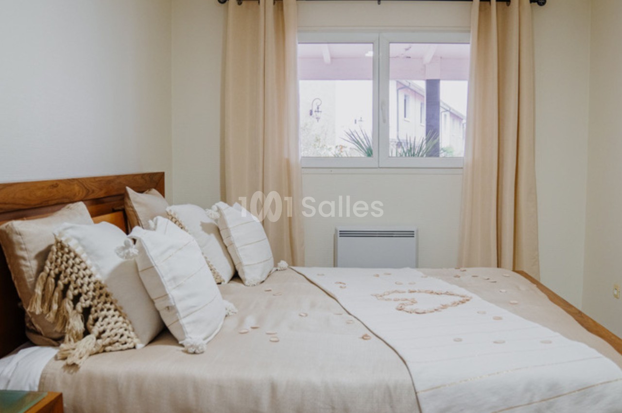 Chambre lumineuse avec lit double, draps beige clair, coussins décoratifs et fenêtre avec rideaux assortis.