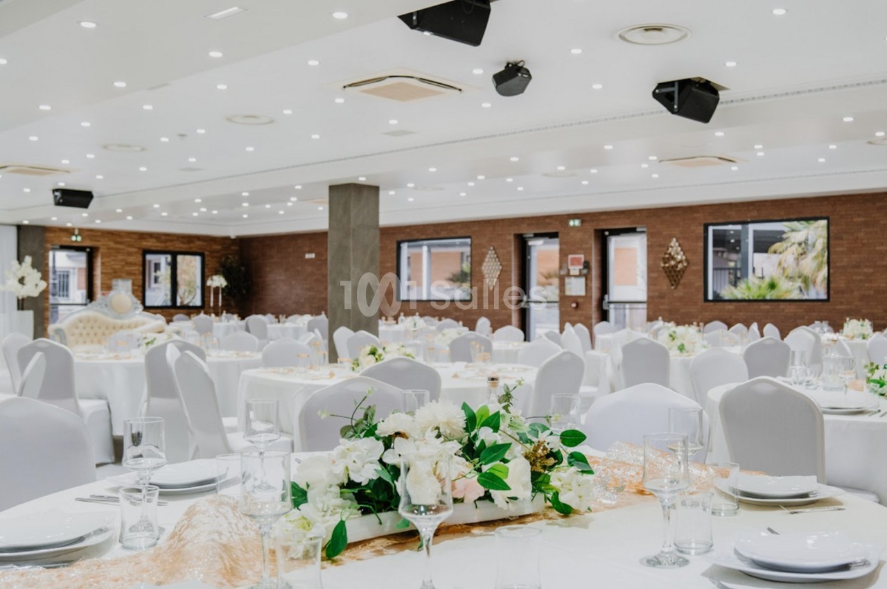 Salle de réception élégante avec tables rondes dressées, nappes blanches et décoration florale au centre.