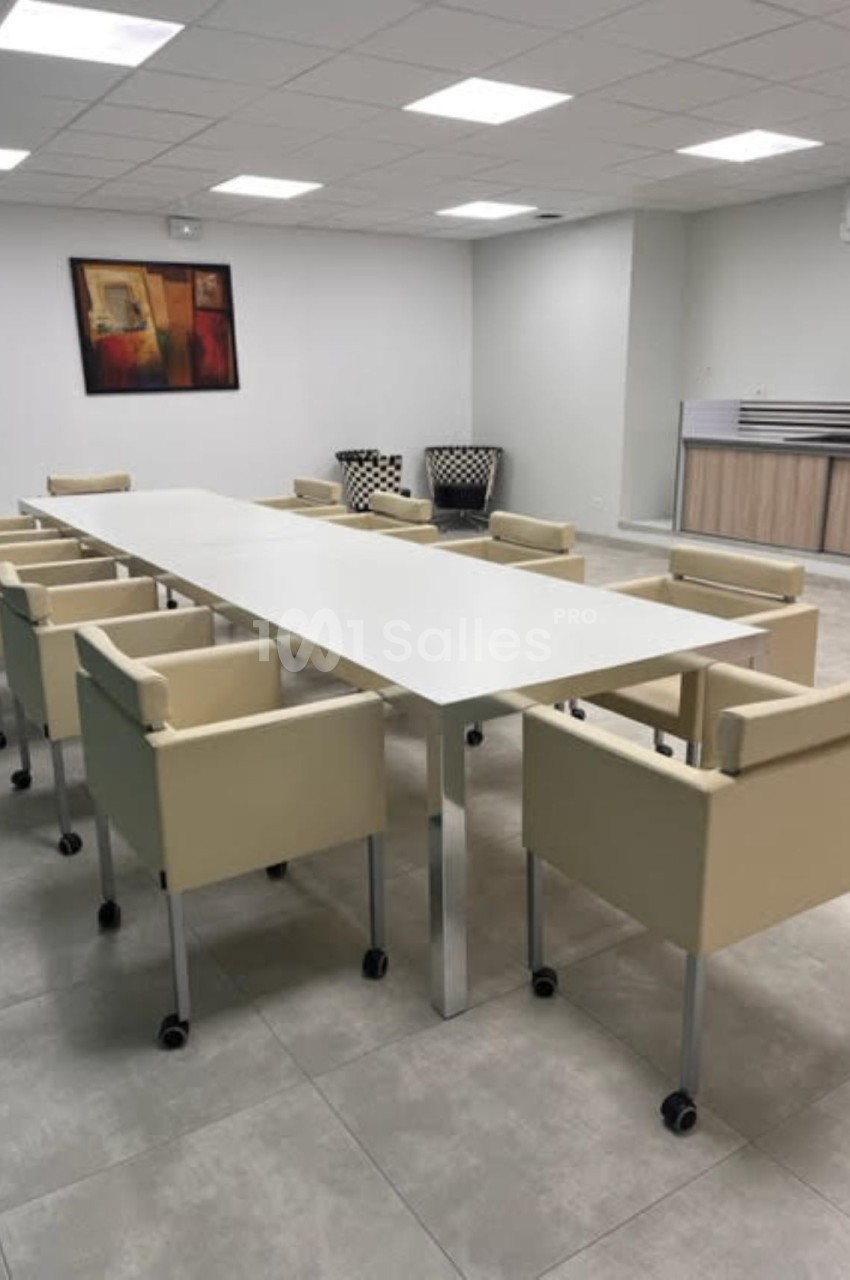 Salle de réunion avec une grande table blanche entourée de chaises beige sur roulettes, et un coin kitchenette en arrière…