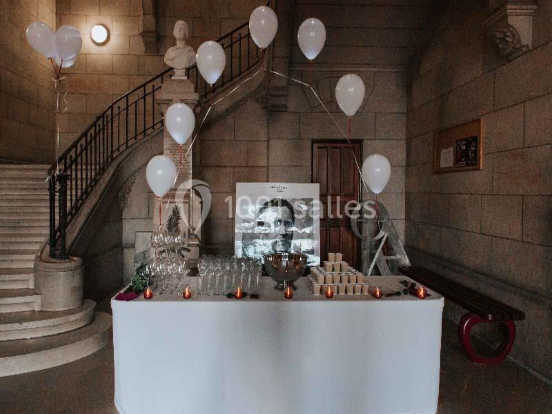 Table décorée avec des ballons blancs, verres, bougies et photo en noir et blanc, dans un hall en pierre avec escalier.