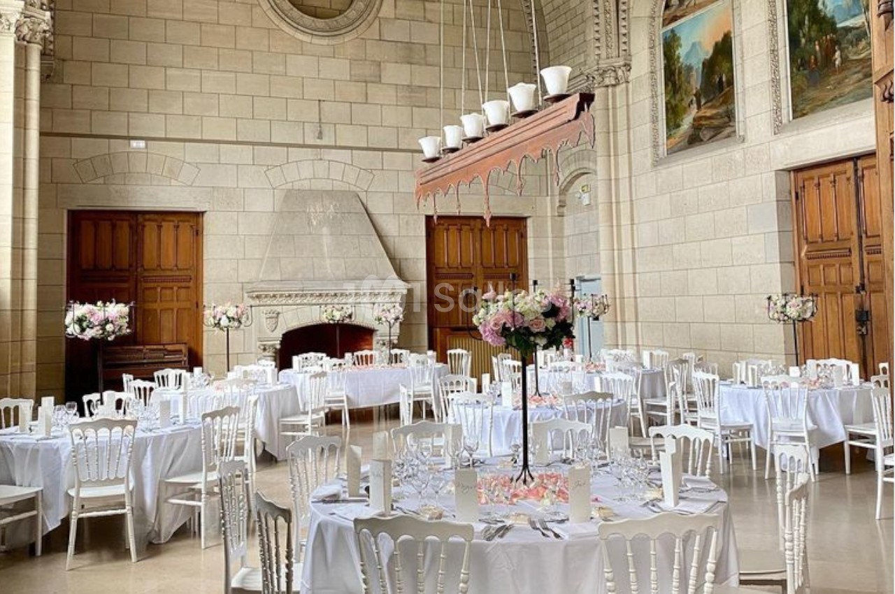 Salle de réception élégante avec tables rondes dressées, chaises blanches et décorations florales raffinées.