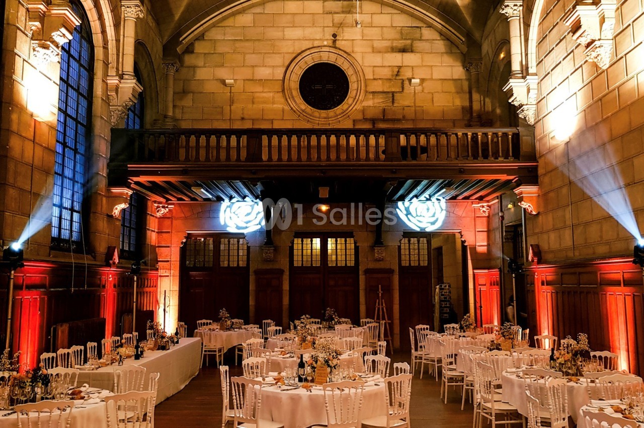 Salle de réception décorée avec des tables dressées, éclairage chaleureux et architecture en pierre avec vitraux.