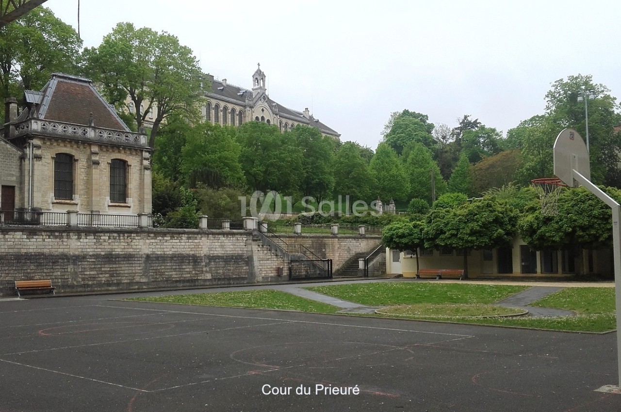Cour extérieure avec terrain de sport, bancs, arbres et bâtiments anciens en arrière-plan.