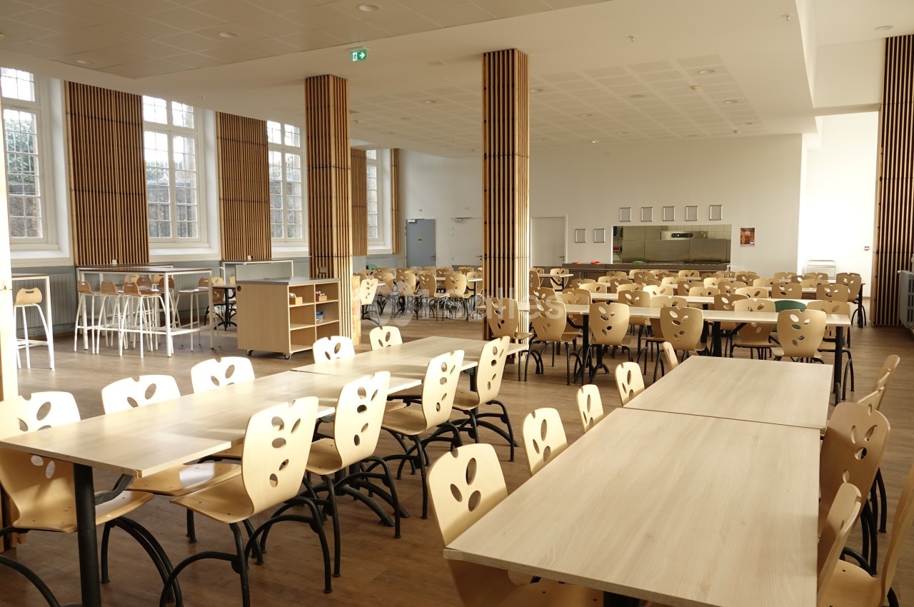Salle lumineuse avec de grandes fenêtres, tables en bois clair et chaises alignées dans un espace de restauration collectif.