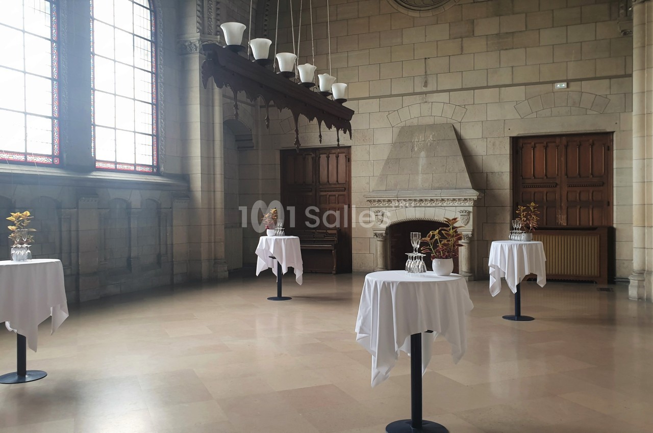 Salle lumineuse avec tables hautes nappées de blanc, décorées de vases contenant des plantes, et cheminée en pierre.