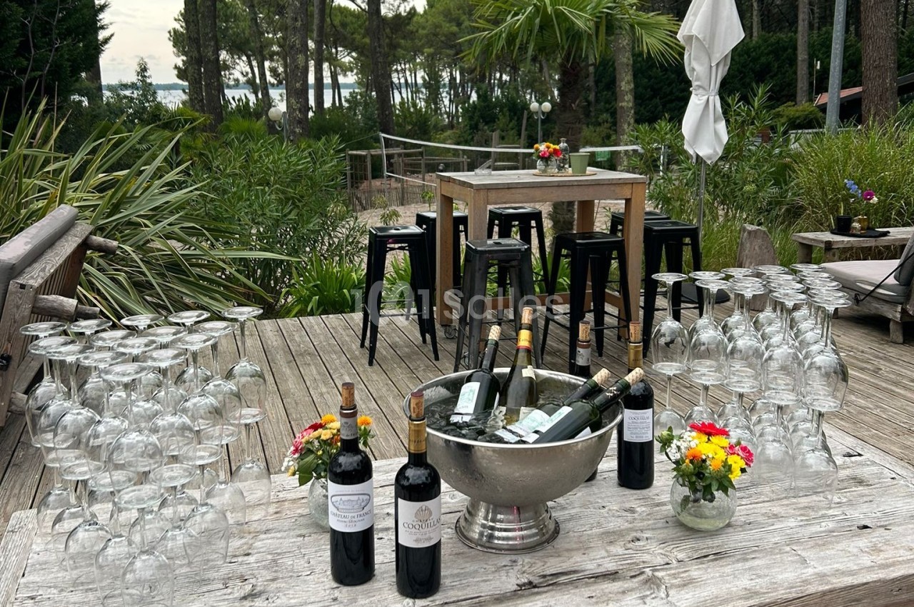 Table en bois avec des bouteilles de vin, des verres empilés et des fleurs, sur une terrasse entourée de végétation.