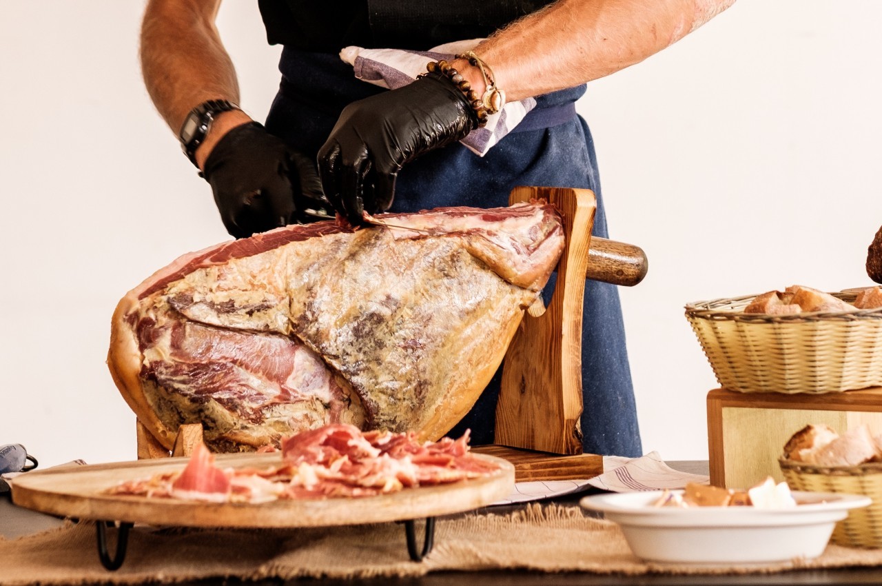 Un artisan découpe un jambon sec sur un support en bois, avec des tranches disposées sur une assiette au premier plan.