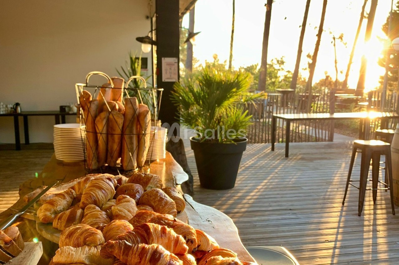Corbeille de baguettes et plateau de croissants sur une table en bois, terrasse ensoleillée en arrière-plan.