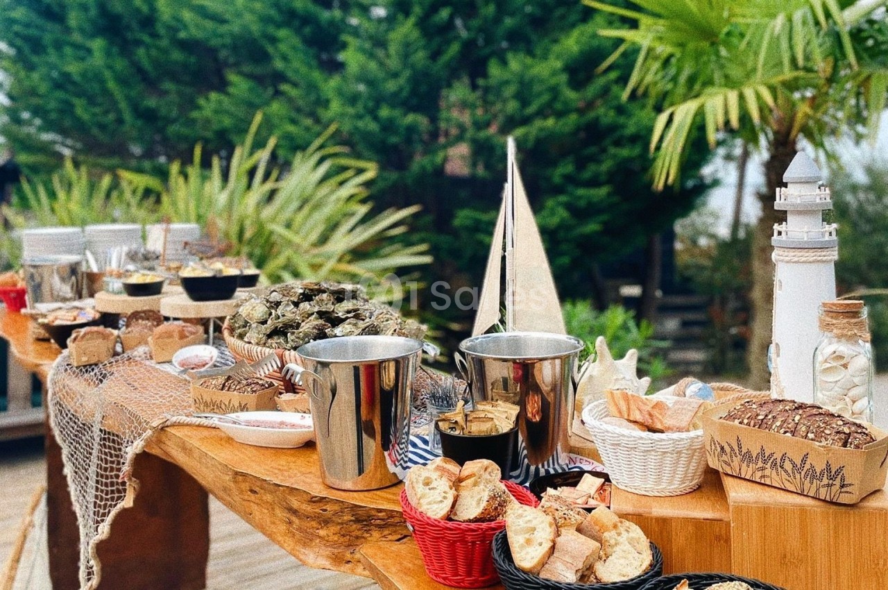 Table en bois garnie de fruits de mer, pains, condiments et décorations marines, entourée de végétation.