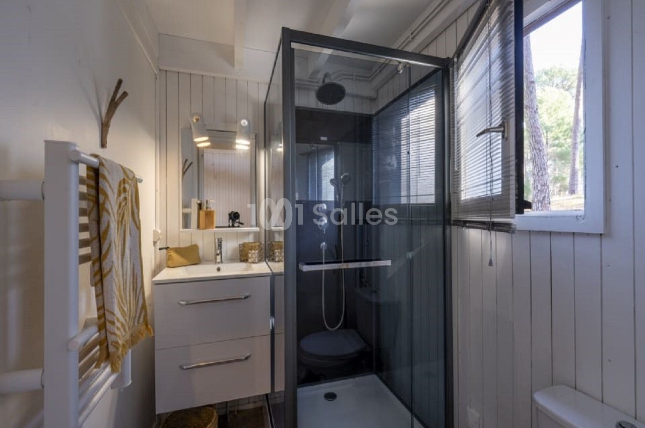 Salle de bain lumineuse avec cabine de douche vitrée, meuble vasque blanc et fenêtre laissant entrer la lumière naturelle.