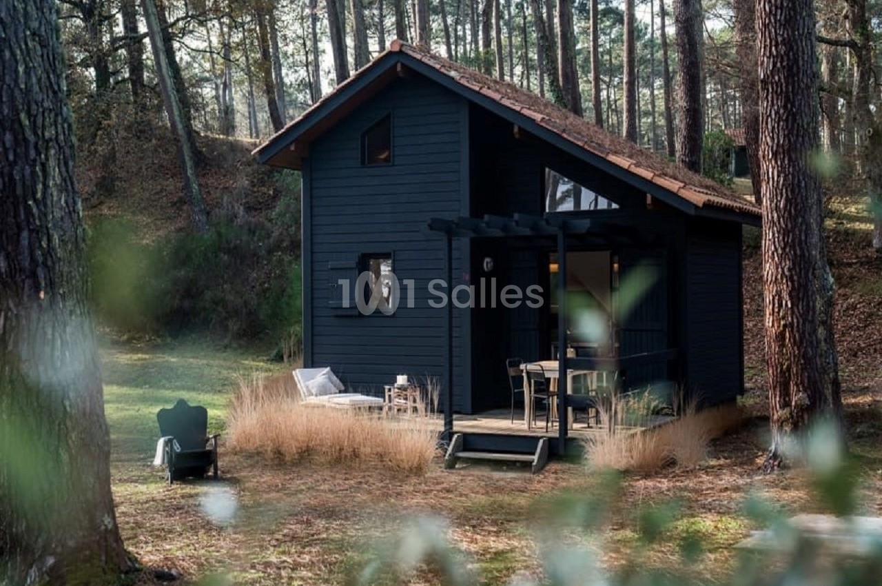 Cabane en bois sombre avec terrasse, entourée de pins et d'herbes hautes dans une clairière forestière.