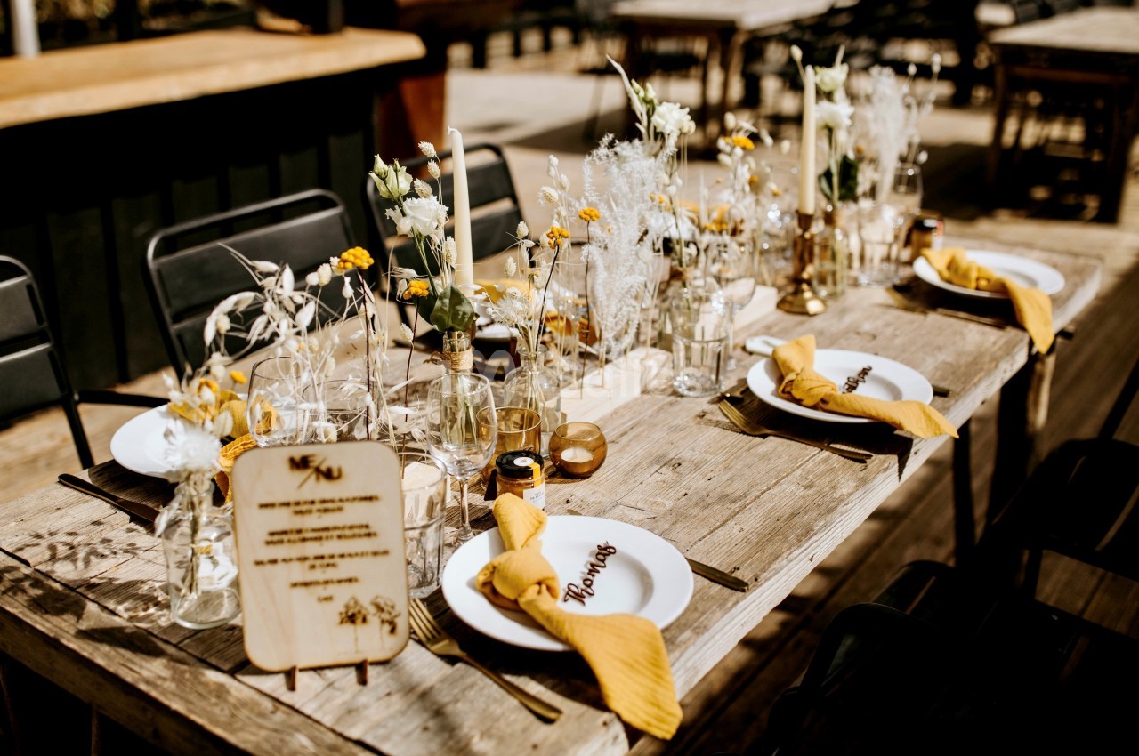 Table en bois décorée pour un repas, avec fleurs, bougies, serviettes jaunes et menus personnalisés.