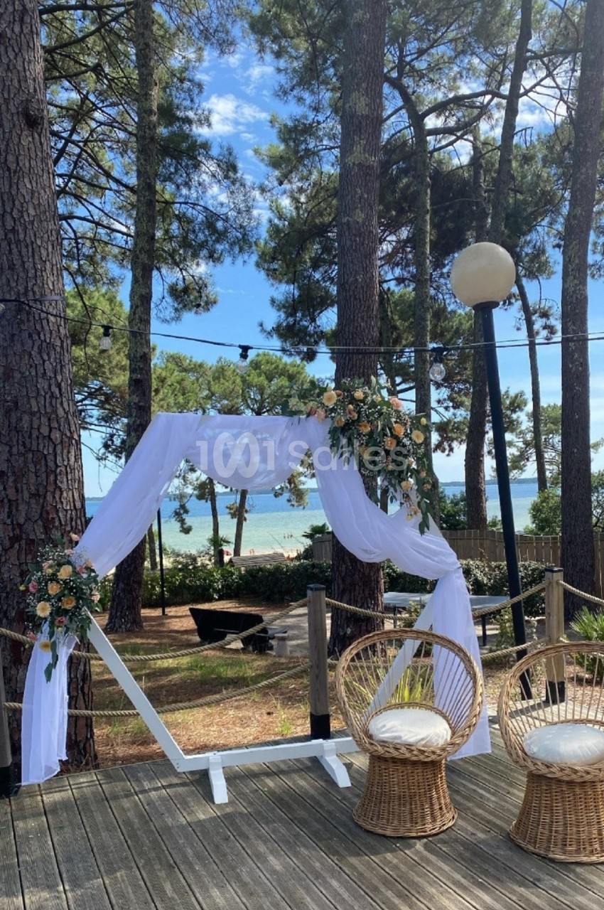 Arche décorée de voilages blancs et de fleurs, entourée de chaises en rotin, avec vue sur une forêt et la mer.