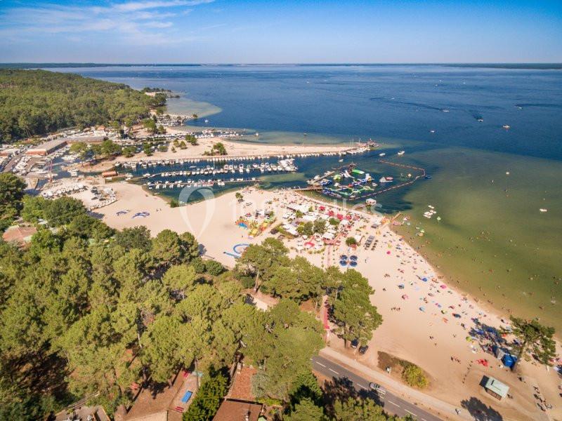 Vue aérienne d'une plage bordée de pins, avec un port de plaisance, des zones de baignade et des activités nautiques.