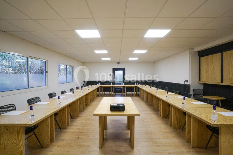 Salle de réunion lumineuse avec tables en U, chaises noires, projecteur central et bouteilles d'eau sur les tables.