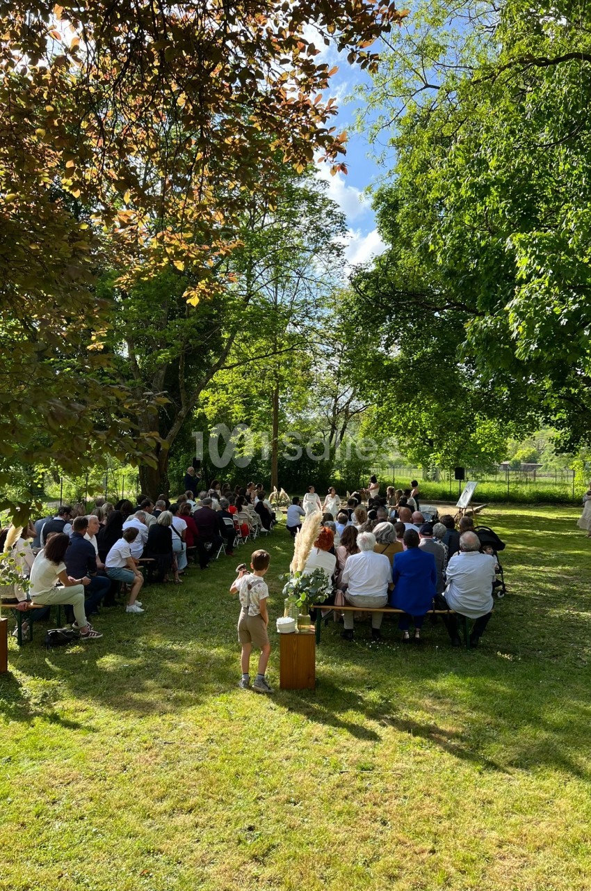 Cérémonie en plein air avec des invités assis sur des bancs dans un jardin verdoyant sous un ciel ensoleillé.