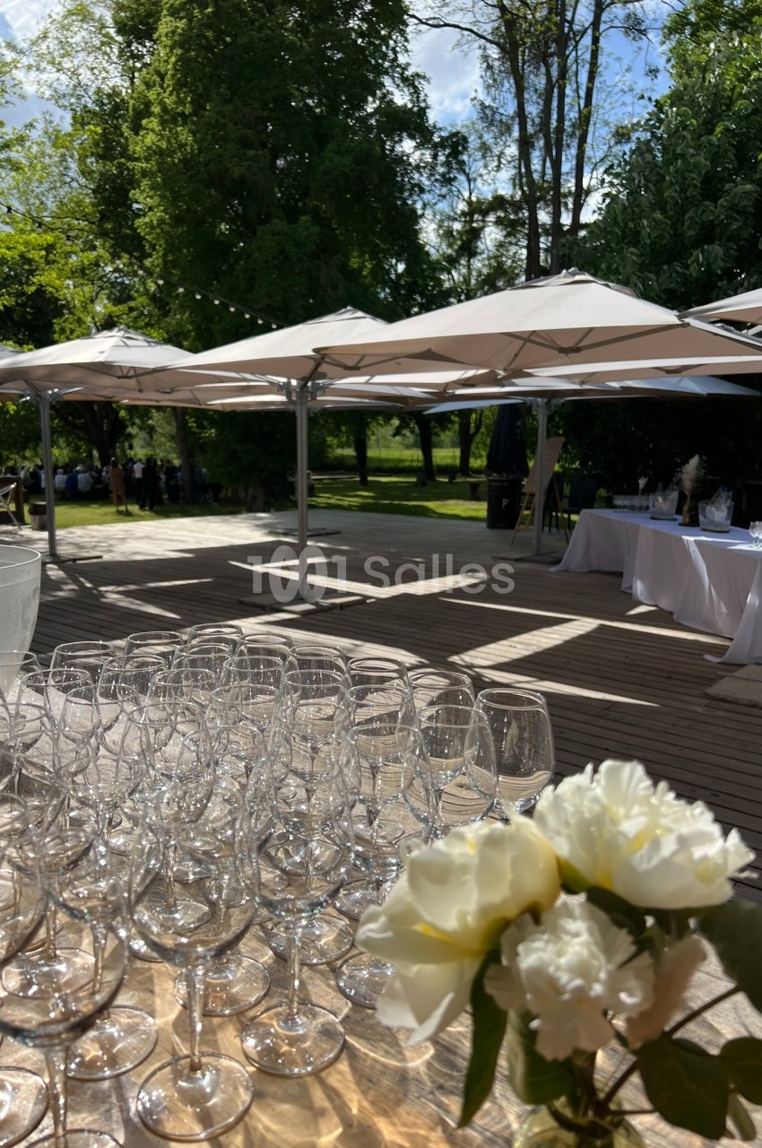 Verres vides alignés sur une table en bois, avec des fleurs blanches au premier plan et des parasols dans un jardin en…