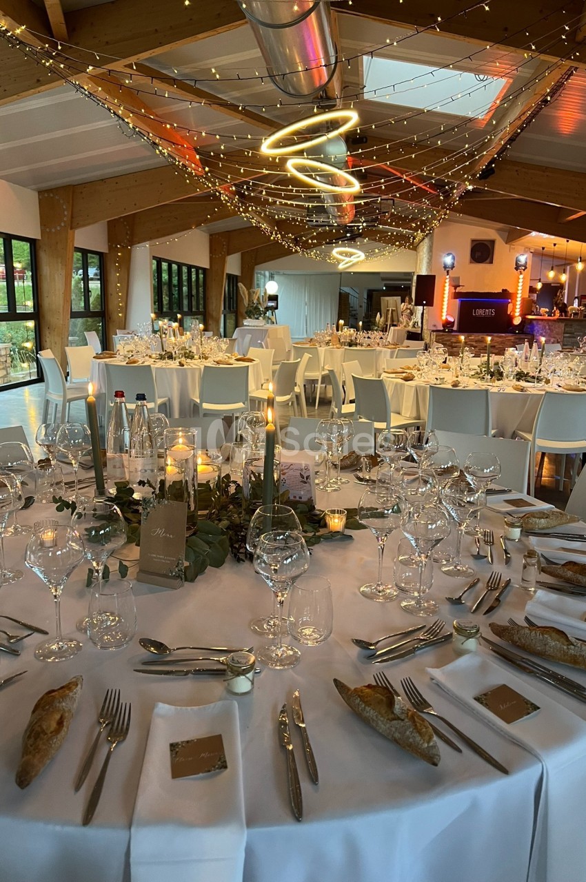 Salle de réception décorée avec des tables rondes dressées, bougies, guirlandes lumineuses et ambiance festive.