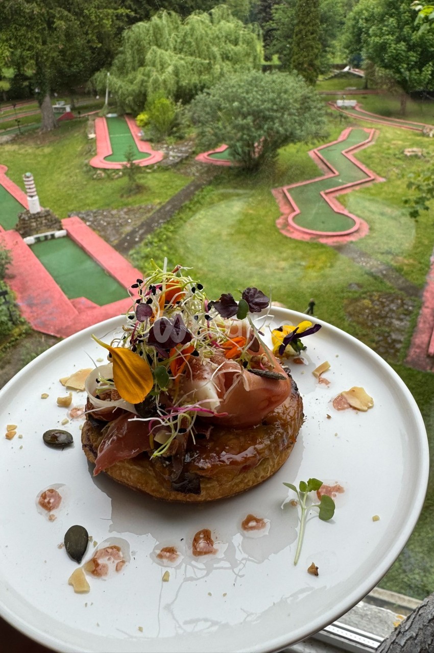 Tarte garnie de légumes, jambon et pousses, présentée devant un mini-golf verdoyant vu depuis une fenêtre.
