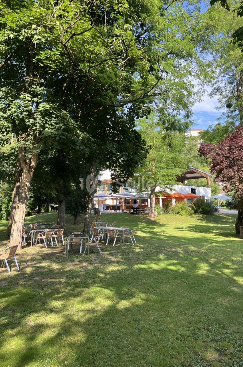 Chaises et tables disposées sur une pelouse ombragée par des arbres, avec un bâtiment en arrière-plan.
