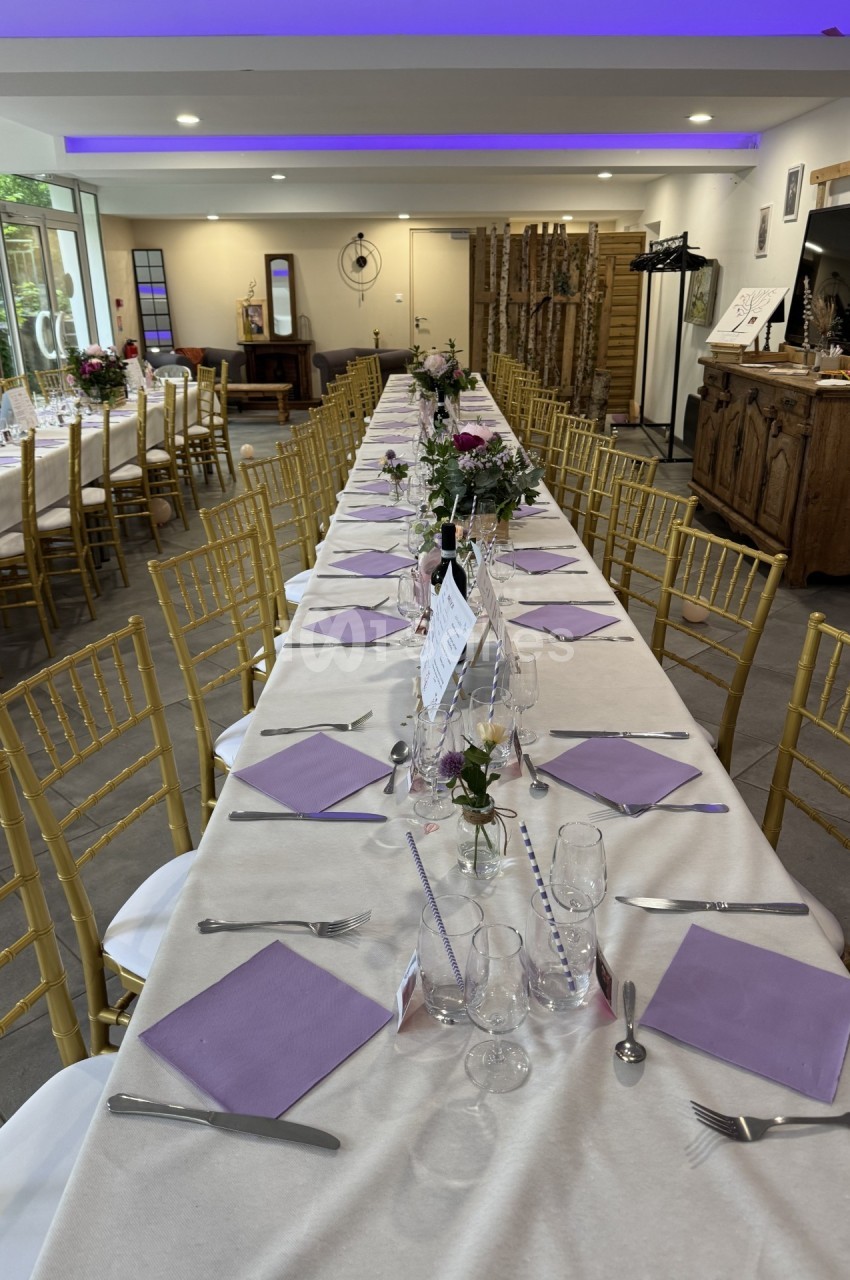 Grande table de réception décorée avec nappes blanches, serviettes violettes, fleurs et chaises dorées dans une salle…