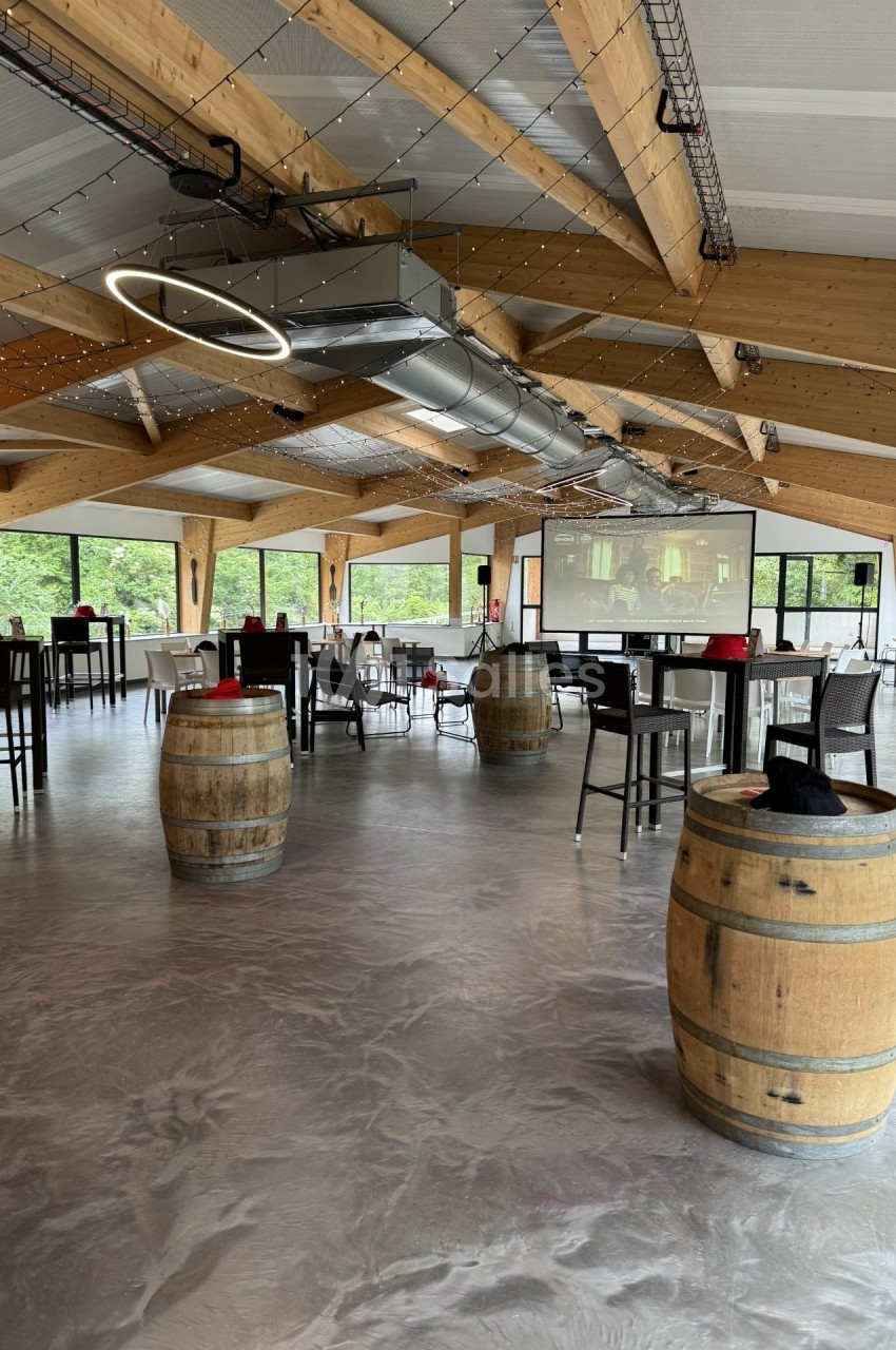 Salle lumineuse avec charpente en bois, tables dressées, tonneaux décoratifs et grandes baies vitrées donnant sur la verdure.