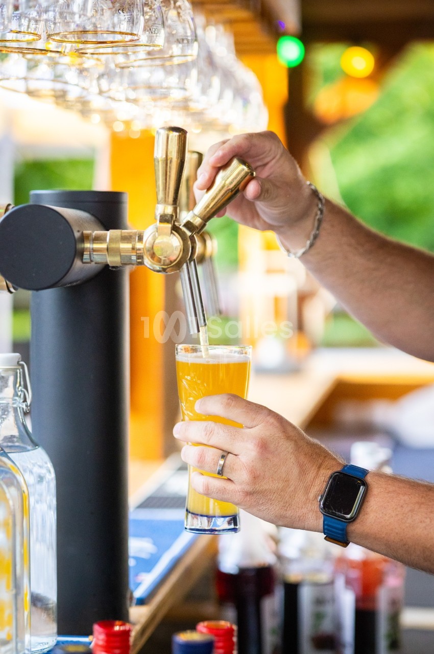 Une main sert une bière blonde à la pression dans un verre, avec un bar en arrière-plan.