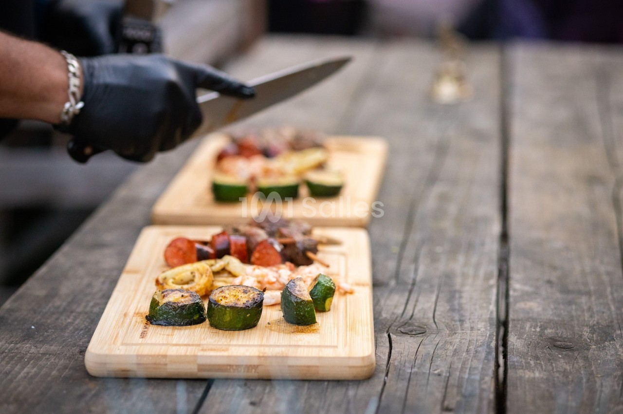 Un cuisinier découpe des aliments sur une planche en bois, avec des légumes grillés et de la viande en arrière-plan.