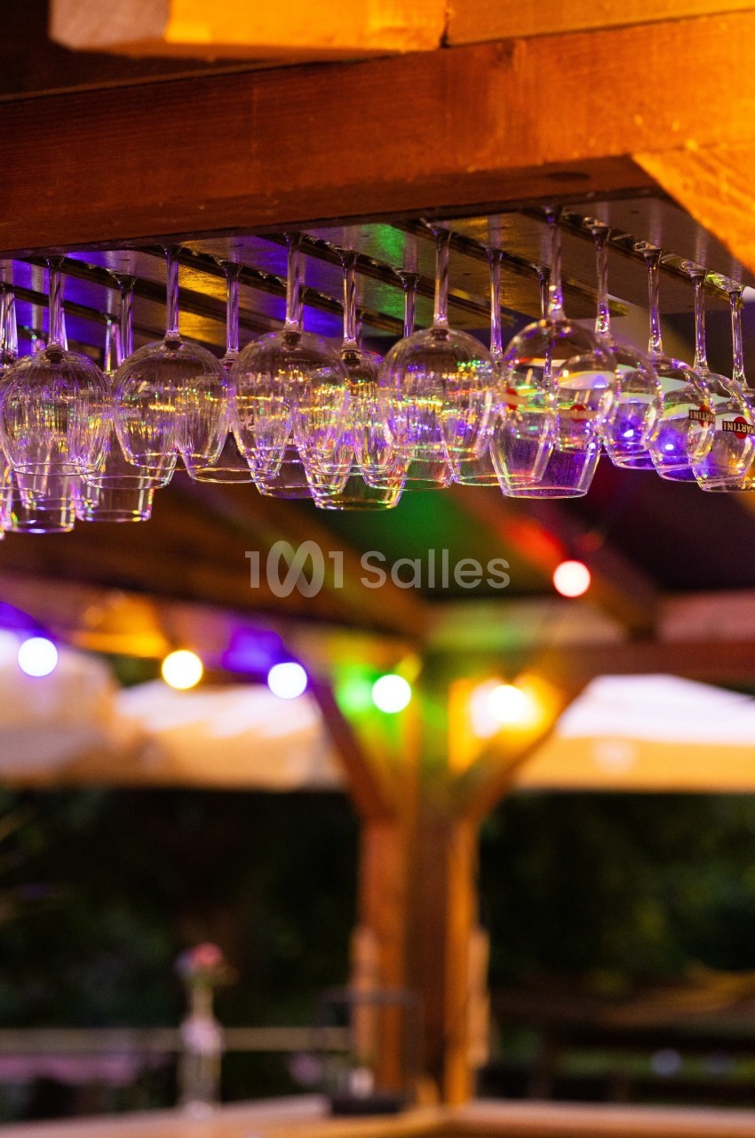 Verres à pied suspendus sous un bar en bois, éclairés par des guirlandes lumineuses colorées en arrière-plan.