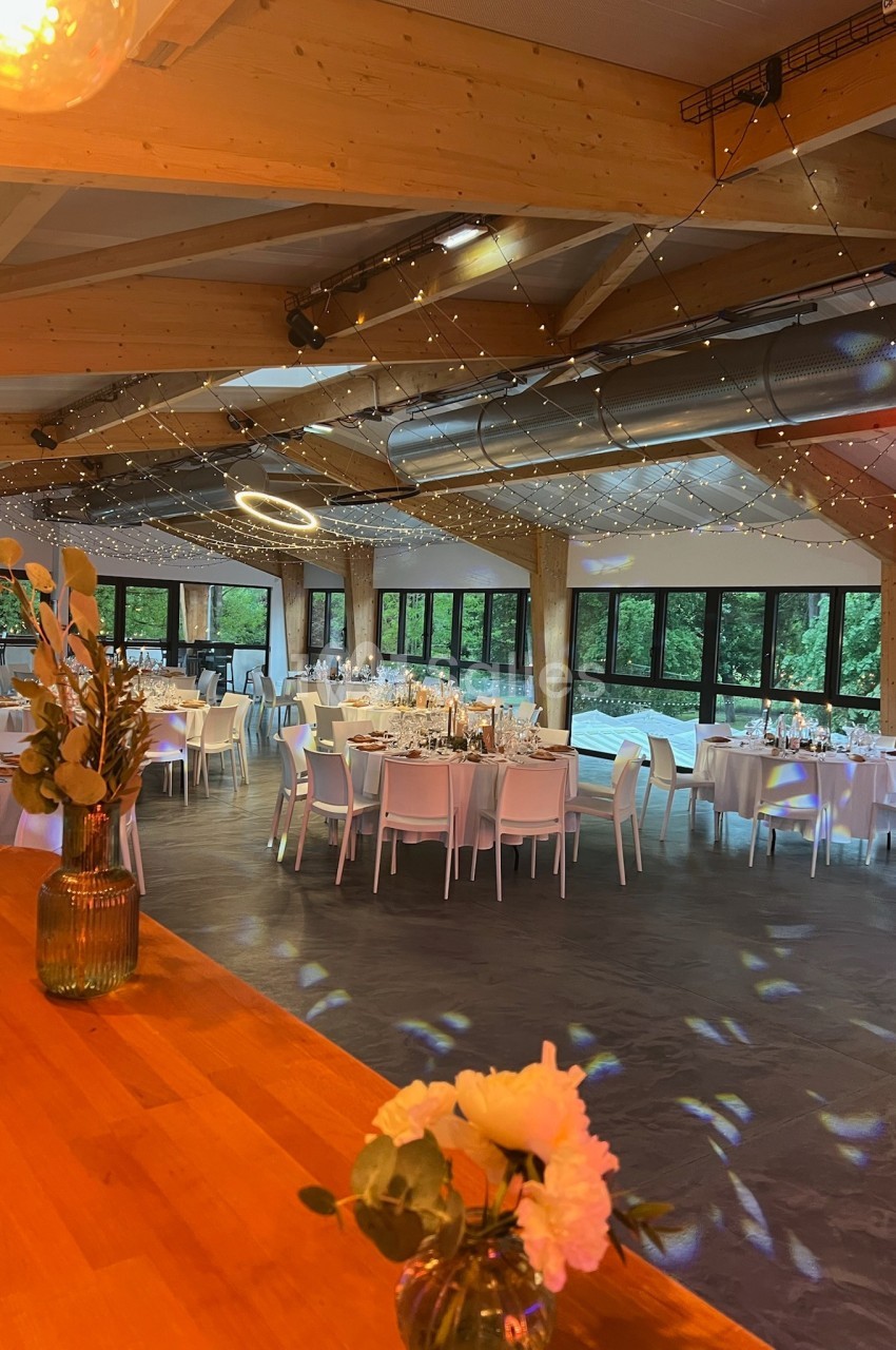 Salle de réception lumineuse avec tables décorées, grandes baies vitrées et plafond en bois orné de guirlandes lumineuses.