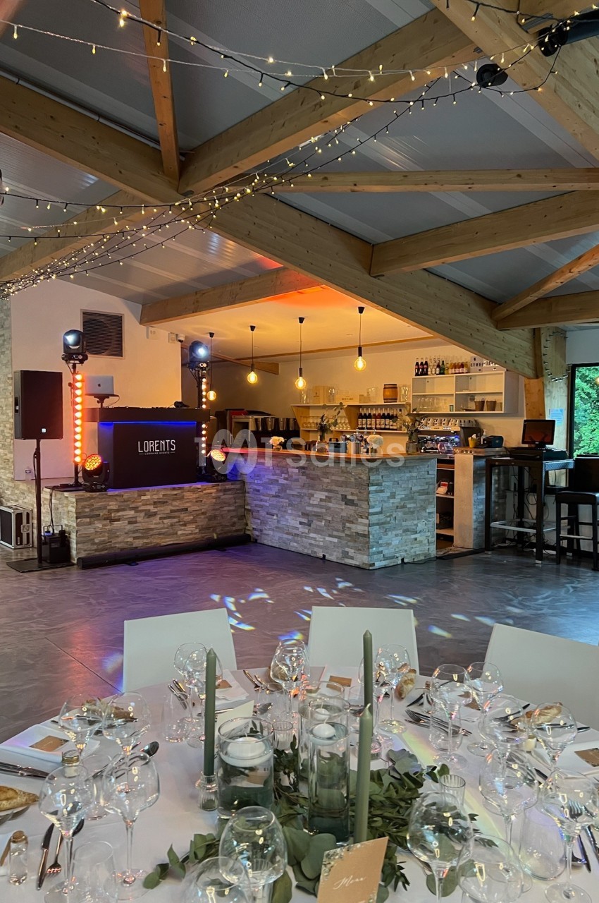 Salle de réception décorée avec des tables dressées, un bar en pierre, des guirlandes lumineuses et un espace DJ.