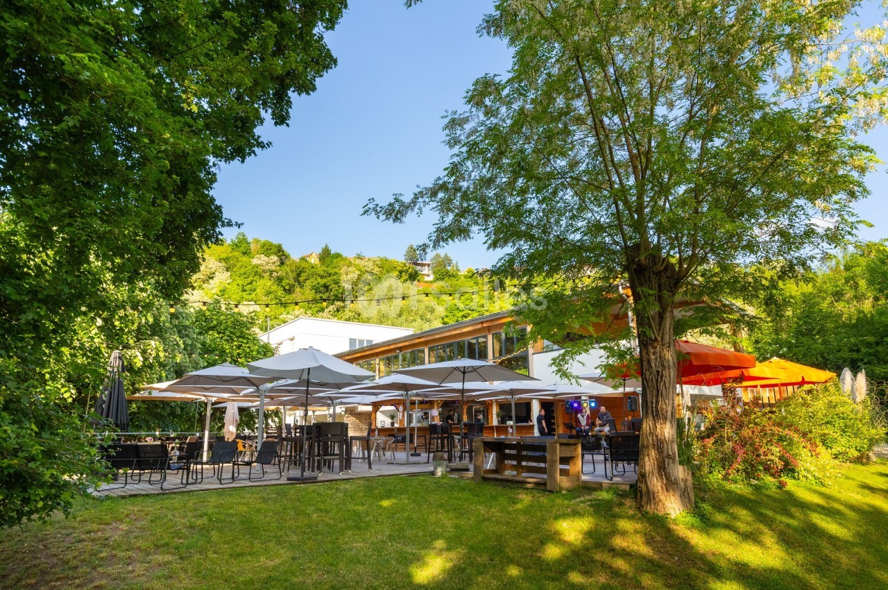 Terrasse d'un restaurant entourée de verdure, avec des parasols blancs et colorés sous un ciel dégagé.