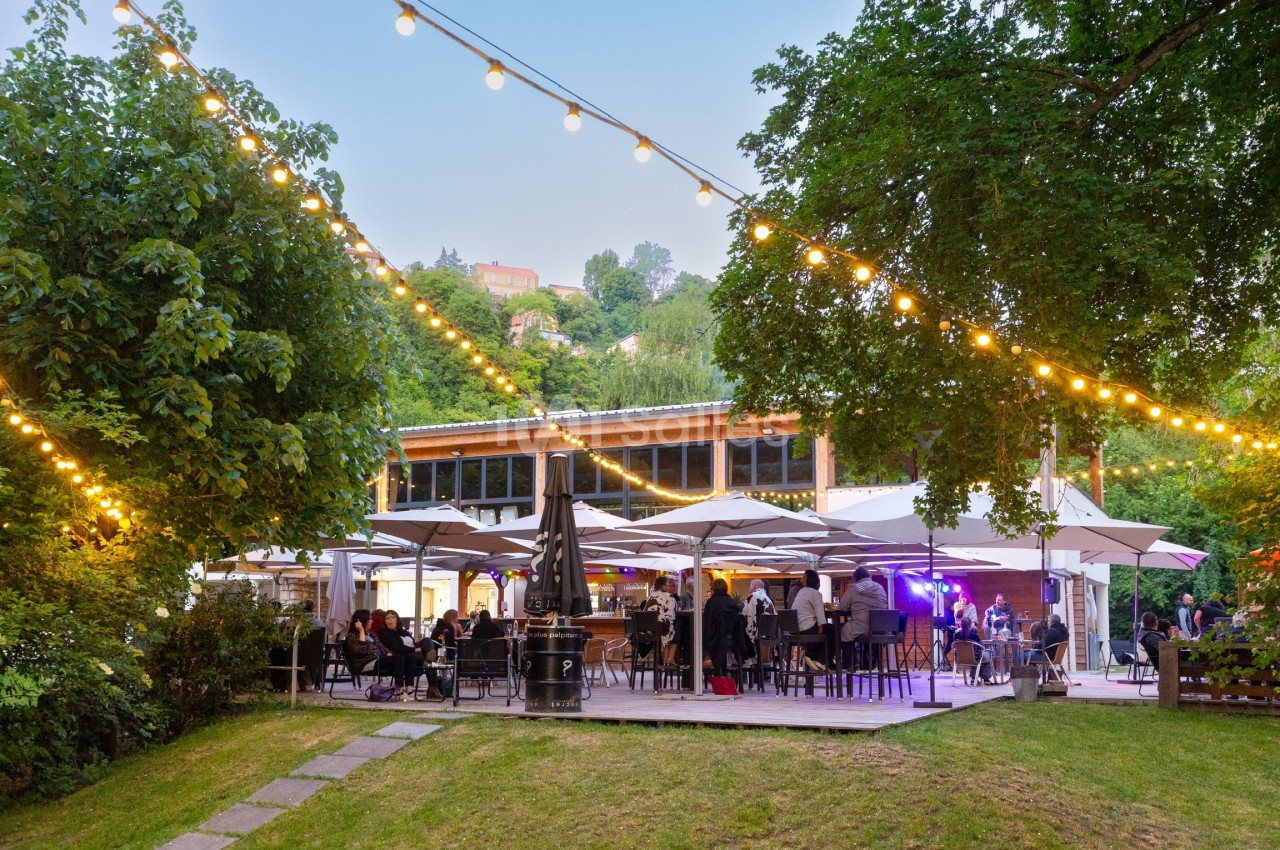 Terrasse d'un restaurant éclairée par des guirlandes lumineuses, entourée de verdure en soirée.