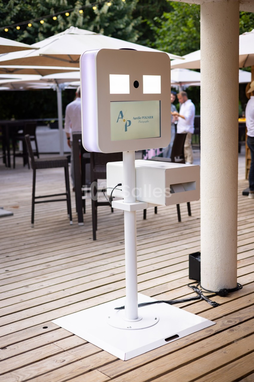 Borne photo interactive blanche installée sur une terrasse en bois, entourée de parasols et de tables hautes.