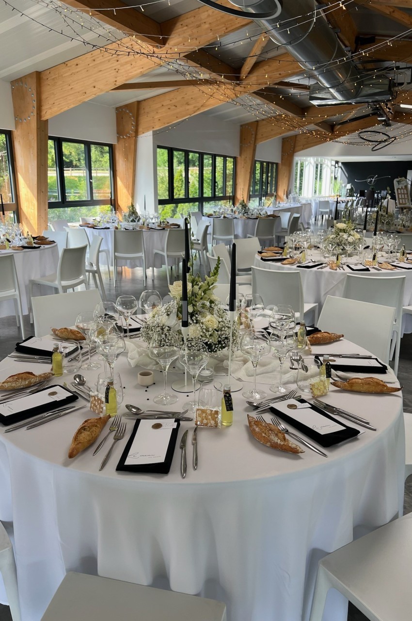Salle de réception lumineuse avec tables rondes dressées, décorations florales blanches et couverts élégants.