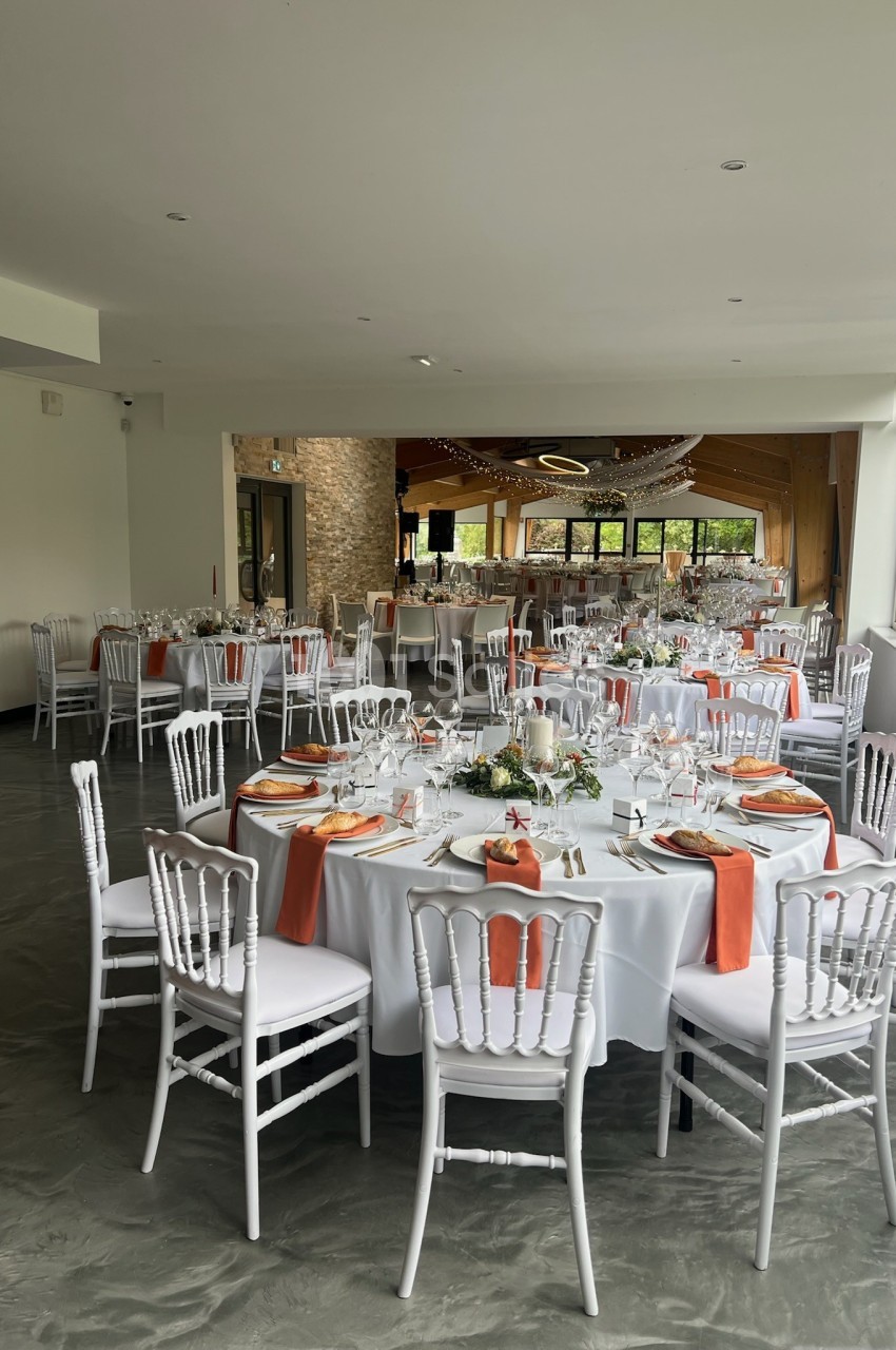 Salle de réception décorée avec des tables rondes, nappes blanches, serviettes orange et chaises blanches.