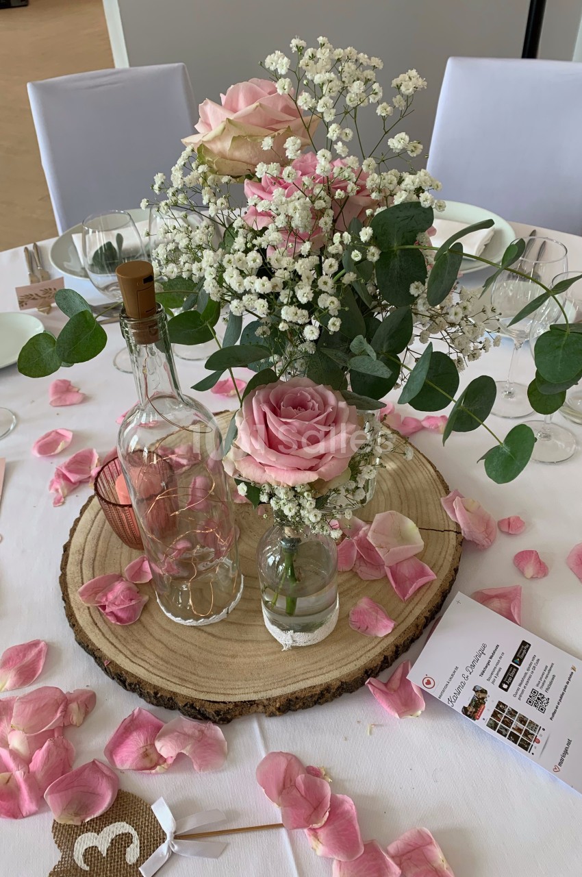 Centre de table avec roses roses, gypsophile, eucalyptus, bouteilles en verre et pétales sur une rondelle de bois.