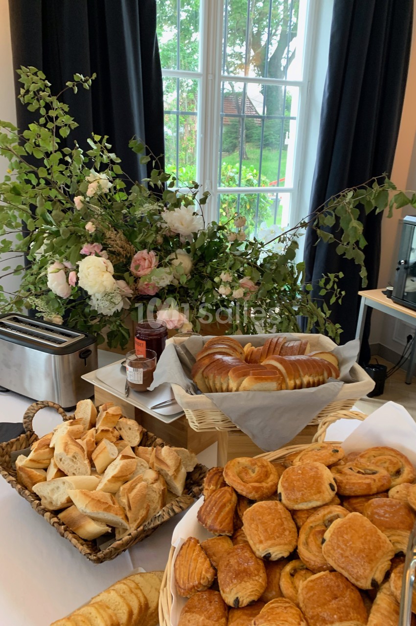 Buffet de petit-déjeuner avec viennoiseries, pain, confiture et grille-pain, devant une fenêtre et des fleurs décoratives.
