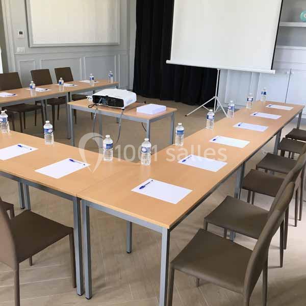 Disposition en U de tables avec chaises, blocs-notes, stylos et bouteilles d'eau, face à un écran de projection.