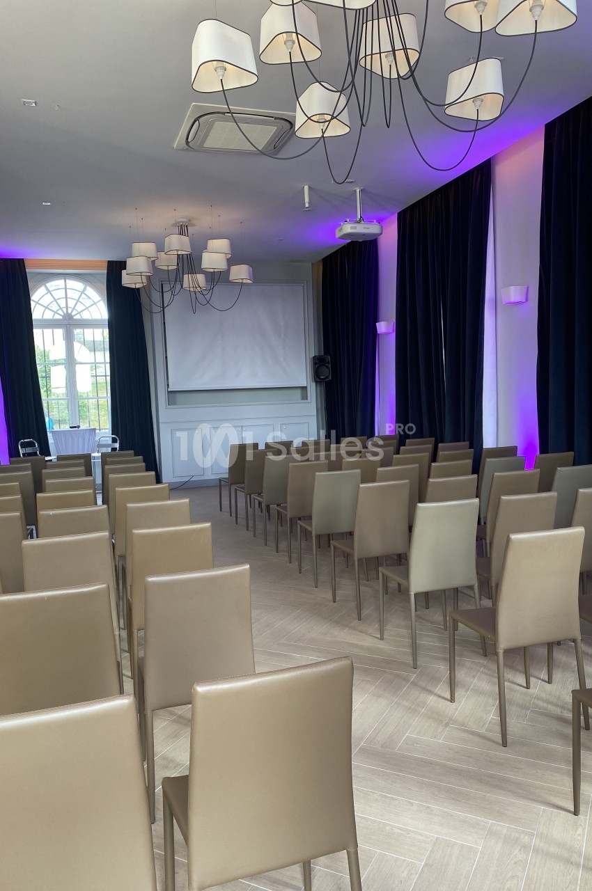 Salle de conférence avec rangées de chaises beige, écran de projection, rideaux sombres et éclairage violet.