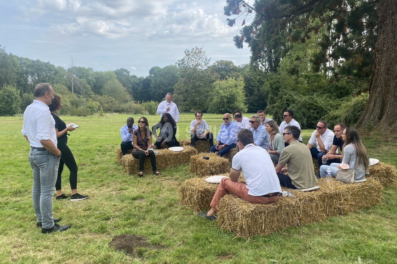 Un groupe de personnes assises sur des bottes de paille en plein air, écoutant deux intervenants debout.