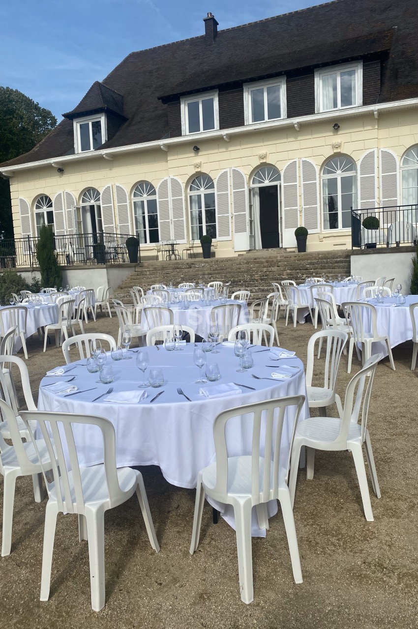 Tables rondes dressées avec nappes blanches et chaises en plastique devant une grande maison de style classique.