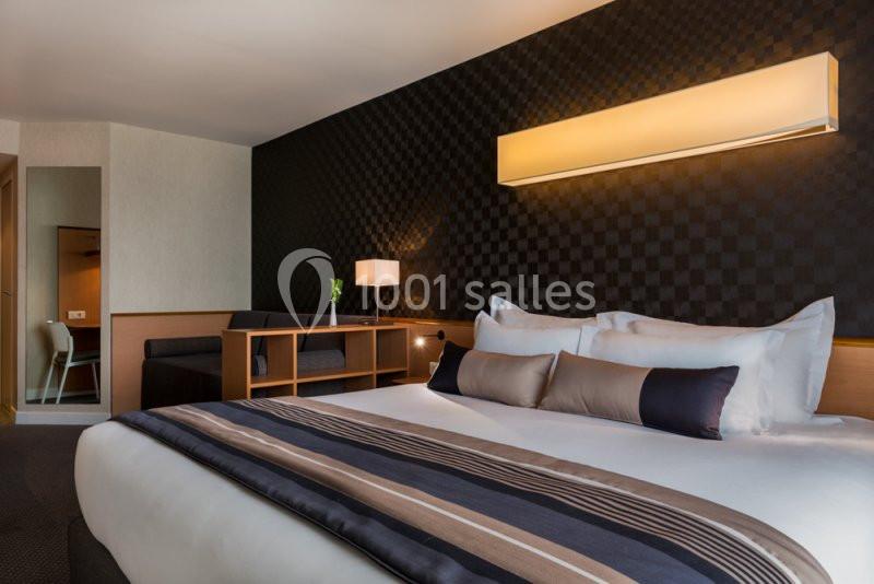 Chambre d'hôtel moderne avec lit double, coussins décoratifs, éclairage mural et coin salon en arrière-plan.