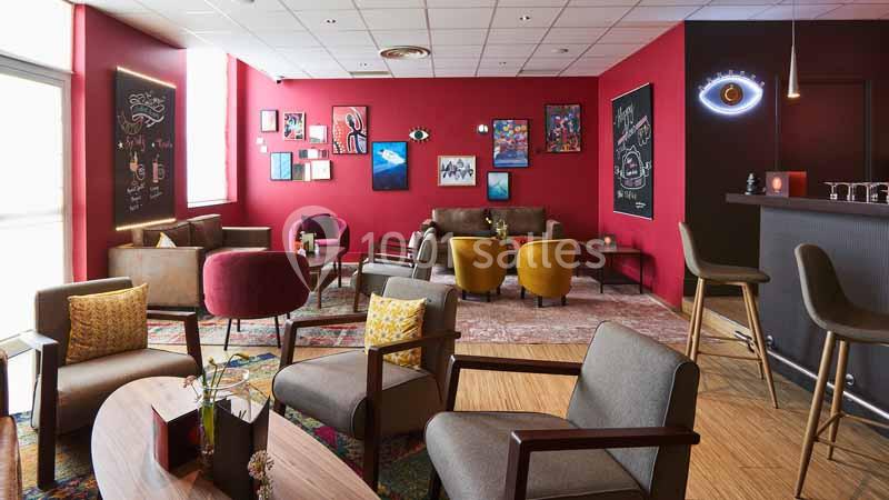 Espace lounge avec fauteuils, tables basses, murs rouges décorés de cadres et tableaux, et un bar à droite.
