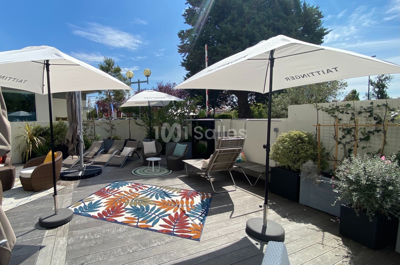 Terrasse ensoleillée avec parasols, chaises longues, tapis coloré et plantes en pots, entourée de verdure.