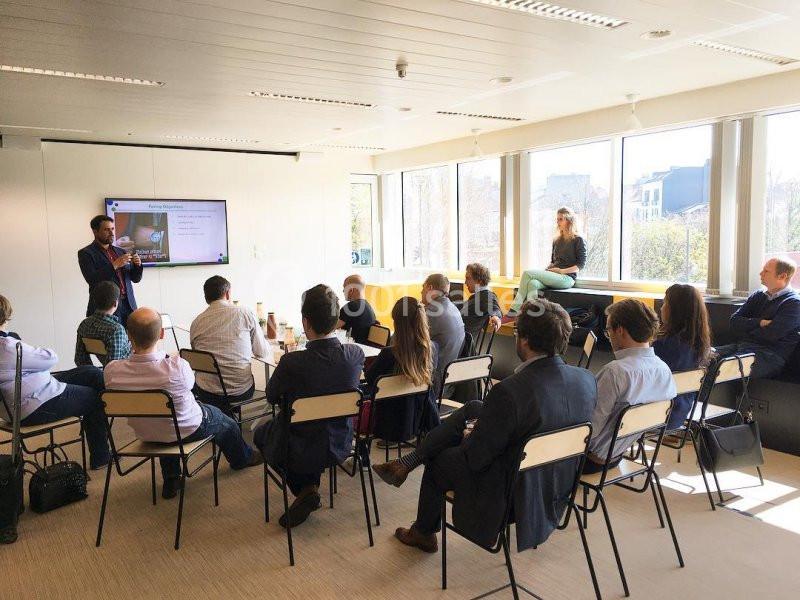 Un homme présente un diaporama à un groupe de personnes assises dans une salle lumineuse avec de grandes fenêtres.